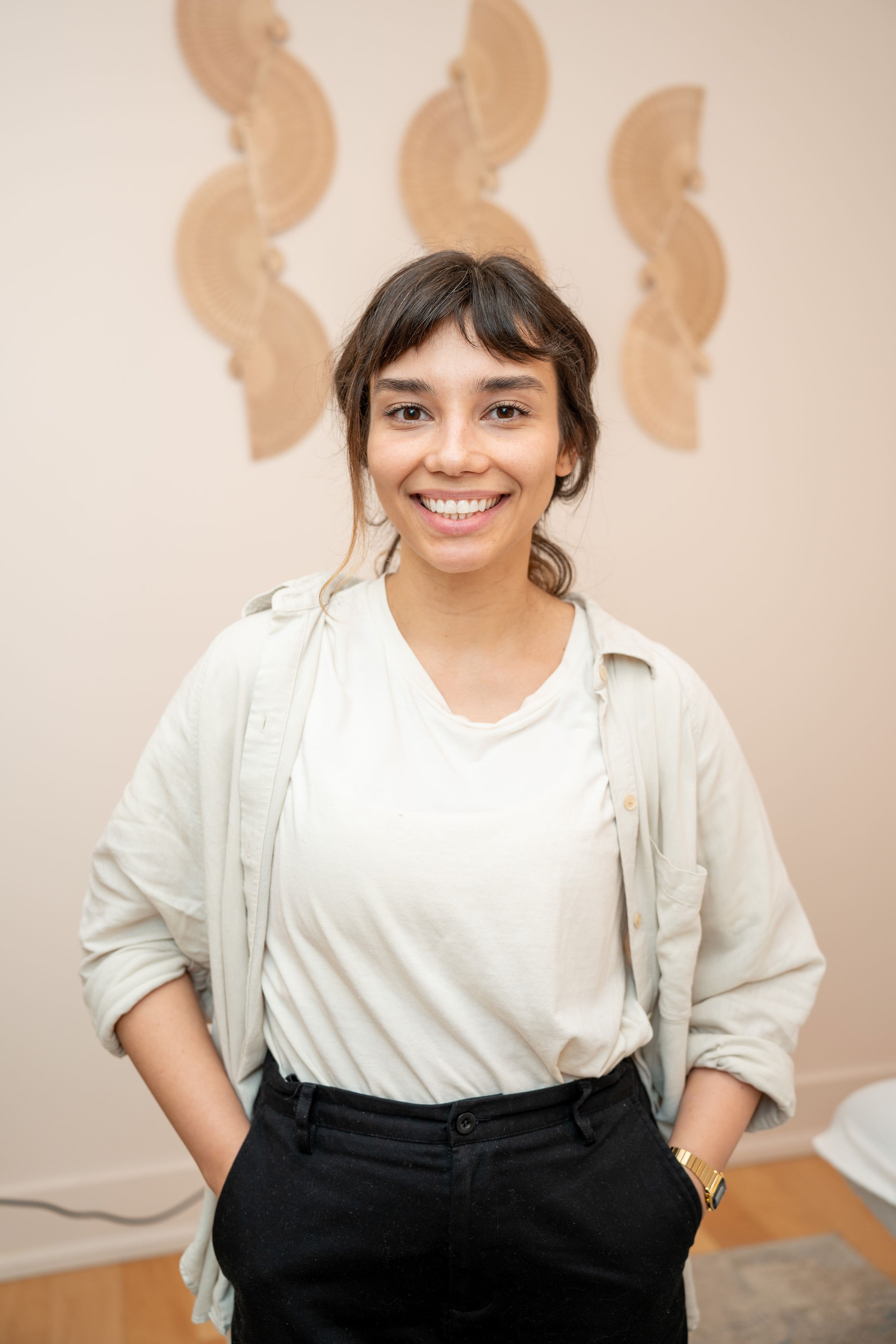Smiling person standing indoors against a decorative wall with abstract art, wearing a light shirt and dark pants.